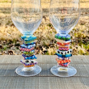 May include: Two clear wine glasses with colorful, stacked, ceramic bases. The bases feature various shapes and patterns in shades of blue, yellow, red, and green. The glasses sit on a wooden surface with a blurred background of greenery.