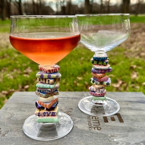 May include: Two clear glass wine glasses with colorful ceramic tiles stacked on the stems. The tiles have various patterns and designs, including the word "LOVE". The glasses are sitting on a wooden surface with a logo.