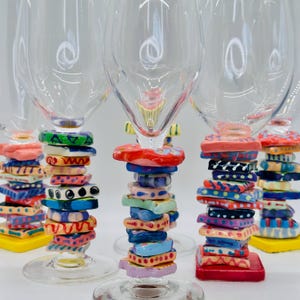 May include: Four clear wine glasses with colorful, patterned ceramic bases stacked on top of the stems. The bases are decorated with geometric shapes, dots, and stripes in a variety of colors, including pink, blue, green, yellow, and red.