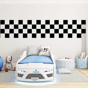 Op de afbeelding: Wit raceauto bedframe met blauw en wit beddengoed, een blauwe zitzak en een knuffelhond. Het bedframe heeft een zwart-wit geruit patroon op het hoofdeinde en de woorden "Police" en "4000 BCD" aan de voorkant.