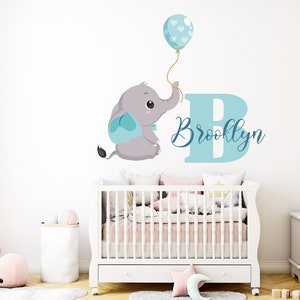 May include: A nursery wall decal featuring a gray cartoon elephant holding a blue balloon with white hearts. A large blue letter "B" and the name "Brooklyn" are also displayed. The decal is above a white crib.