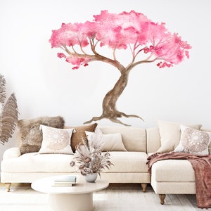 May include: A white sectional sofa with a pink and brown watercolor tree decal on the wall behind it. The sofa has a throw blanket and pillows. There is a vase with dried pampas grass and a coffee table with books and a vase with dried flowers.