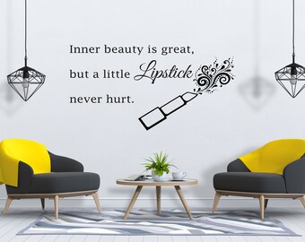 Può includere: Un adesivo da parete in bianco e nero con la citazione "La bellezza interiore è grandiosa, ma un po' di rossetto non ha mai fatto male." La citazione è accompagnata da un'immagine stilizzata di un tubetto di rossetto con un tocco di volute decorative.