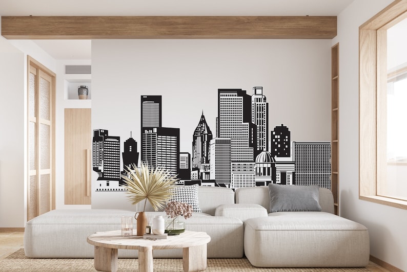 May include: A black and white cityscape wall decal featuring a skyline of tall buildings. The decal is applied to a white wall in a modern living room with a light grey sectional sofa and a wooden coffee table.