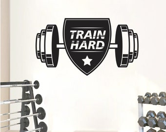 May include: Black and white wall decal featuring a barbell with weights on each side and a shield with the text "TRAIN HARD" and a star in the center.
