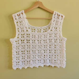 May include: A white crochet top with a square neckline and a scalloped hemline. The sleeveless top is made of a delicate, open-weave pattern, and is hanging on a wooden hanger against a yellow background. The top is a handmade garment.