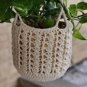 Könnte beinhalten: Ein cremefarbener gehäkelter Hängekorb mit einer grünen Pflanze mit Blättern.