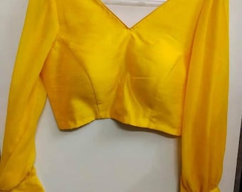 Può includere: Una camicetta giallo brillante a maniche lunghe con un profondo scollo a V, spalle strutturate e polsini aderenti ai polsi. Il capo è realizzato in un tessuto liscio di colore solido ed è esposto su una gruccia di plastica trasparente.