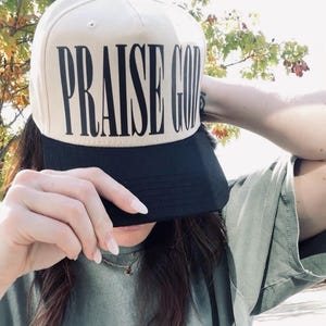 May include: A beige and black baseball cap with the words "PRAISE GOD" in large, bold, black letters across the front. The cap has a black brim and a black button on top. The background is blurred, with green foliage.