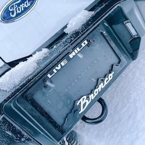 May include: A close-up of a black license plate frame with the words "Live Wild" and "Bronco" on it. The frame is attached to a white Ford Bronco with a Ford logo on the back.