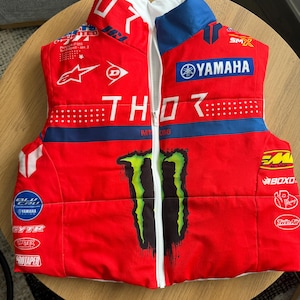 May include: A red sleeveless puffer vest featuring a white front zipper and a large green Monster Energy logo. The vest is covered in various white and blue racing brand logos including Yamaha, Thor, Alpinestars, Dunlop, and ProTaper.