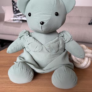 May include: A sage green stuffed bear toy featuring a smooth fabric exterior, round ears, and small black bead eyes and nose. The bear is wearing a matching sage green dress with ruffled cap sleeves and a gathered bodice detail on the front.