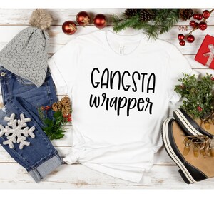 Könnte beinhalten: Weißes T-Shirt mit dem Text "Gangsta Wrapper" in schwarzen Buchstaben. Das Shirt liegt auf einer weißen Holzoberfläche mit einer grauen Mütze, blauen Jeans, einer silbernen Schneeflocke, einer roten Geschenkbox und grünen Kiefernästen mit roten Beeren.