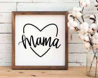 May include: A white wooden sign with a brown frame, featuring a black heart with the word "Mama" inside. The sign is sitting on a wooden surface.