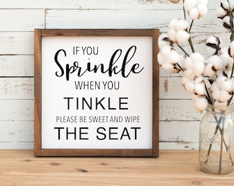 May include: A framed wooden sign with the text "If you Sprinkle when you Tinkle Please be sweet and wipe the seat" in black script. The sign is on a wooden surface with a mason jar of cotton stems.
