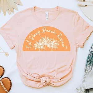 May include: A pink t-shirt with the text "Eat Sleep Beach Repeat" in a white semi-circle with palm trees inside. The text is in a handwritten font.