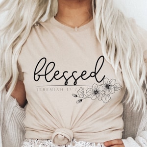 Könnte beinhalten: Ein beiges T-Shirt mit dem Wort "blessed" in schwarzer Kursivschrift und dem Text "Jeremiah 17:7" darunter. Das Shirt hat ein florales Design mit schwarzen Umrissen.
