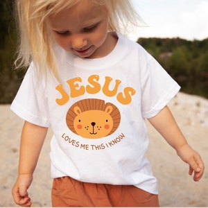 Puede incluir: Una camiseta blanca con un gr&aacute;fico de un rostro de le&oacute;n y el texto "JESUS LOVES ME THIS I KNOW".