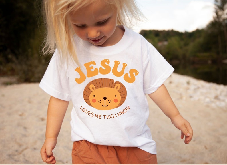 Puede incluir: Una camiseta blanca con un gr&aacute;fico de un rostro de le&oacute;n y el texto "JESUS LOVES ME THIS I KNOW".