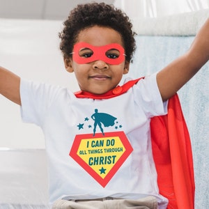 Puede incluir: Un ni&ntilde;o peque&ntilde;o con una camiseta blanca, una capa roja y una m&aacute;scara de superh&eacute;roe roja. La camiseta tiene un logotipo de superh&eacute;roe rojo y amarillo con el texto "Puedo hacer todas las cosas por medio de Cristo".