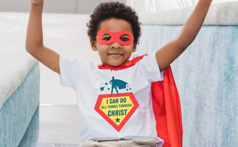 Puede incluir: Un ni&ntilde;o peque&ntilde;o con una camiseta blanca, una capa roja y una m&aacute;scara de superh&eacute;roe roja. La camiseta tiene un logotipo de superh&eacute;roe rojo y amarillo con el texto "Puedo hacer todas las cosas por medio de Cristo".