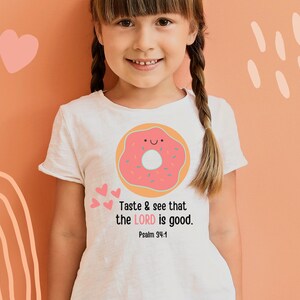 Puede incluir: Una camiseta blanca con un gr&aacute;fico de donut rosa y el texto "Taste & see that the LORD is good. Psalm 34:8" impreso en ella.