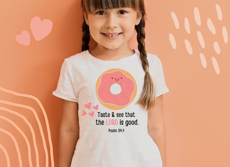 Puede incluir: Una camiseta blanca con un gr&aacute;fico de donut rosa y el texto "Taste & see that the LORD is good. Psalm 34:8" impreso en ella.