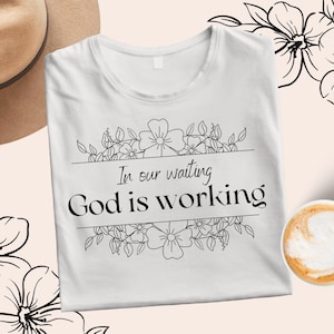Pode incluir: Camiseta branca com um design floral e o texto "In our waiting God is working".