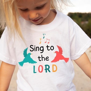 Puede incluir: Camiseta blanca con un dise&ntilde;o gr&aacute;fico colorido con dos p&aacute;jaros y el texto "Sing to the LORD".