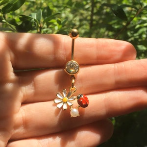 May include: A gold-toned navel ring featuring a clear round gemstone at the top, with a dangling charm consisting of a white daisy with a yellow center, a small red and black ladybug, and a single white pearl-like bead at the bottom.