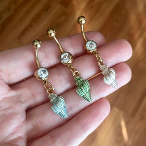May include: Three gold-coloured belly button rings with clear gem accents and seashell charms in blue, green, and silver. The curved bars have a small ball at the top and a larger gem at the centre.