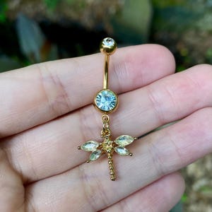 Puede incluir: Anillo para el ombligo de color dorado con un dije de libélula. El anillo presenta una gema clara y redonda en la parte superior y una gema azul claro más grande en el centro. El dije de libélula tiene gemas claras en forma ovalada en sus alas.