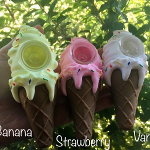 Può includere: Tre oggetti a forma di cono gelato in giallo, rosa e bianco, ciascuno con una base a cono marrone. Quello giallo è etichettato "Banana", quello rosa "Strawberry" e quello bianco "Vanilla". Ognuno ha una caratteristica circolare trasparente.