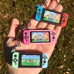 May include: Three colorful video game console keychains. The keychains are in blue and red, pink, and green and blue. Each keychain has a small screen displaying a colorful scene. The keychains are attached to silver keyrings.