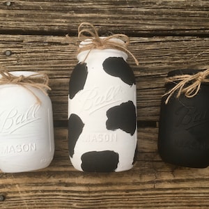 Puede incluir: Tres tarros Ball Mason pintados con detalles de cuerda. Un tarro es blanco, otro es blanco con manchas negras de vaca y el tercero es negro. Los tarros están dispuestos sobre una superficie de madera rústica.