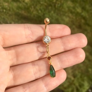 Puede incluir: Un piercing para el ombligo de color dorado con un diseño de barra curva. La bola superior y el dije principal presentan una gema grande, clara y redonda. Una gema roja más pequeña y una gema verde en forma de lágrima cuelgan debajo.