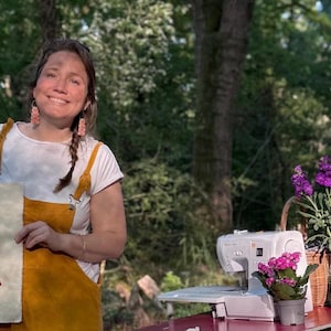 May include: A woman wearing a white shirt and yellow dungarees is holding a piece of fabric with an embroidered portrait of a person with colourful hair. The portrait is on a white background. The woman is standing in a wooded area with a sewing machine and flowers in the background.