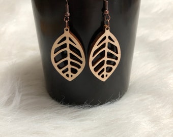 Puede incluir: Un par de pendientes de madera con forma de hoja con un diseño delicado e intrincado. Los pendientes son de color marrón claro y tienen ganchos de color cobre.