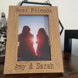 May include: A wooden photo frame with the text "Best Friends" and "Amy & Sarah" engraved on the top and bottom. The frame holds a photo of two people standing on a beach at sunset.