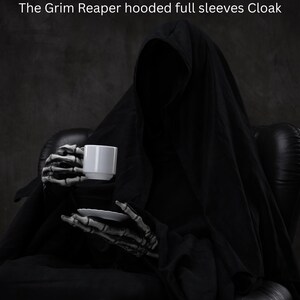 May include: A black hooded cloak with full sleeves worn by a figure with skeletal hands holding a white ceramic coffee cup and saucer. The text at the top of the image reads: The Grim Reaper hooded full sleeves Cloak. The garment is solid black.