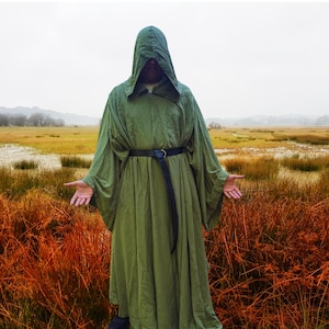 May include: A floor-length olive green hooded robe with long, wide sleeves and a black leather belt featuring a metal ring buckle. The garment is loose-fitting, made of a solid-coloured fabric, and covers the torso and arms completely.