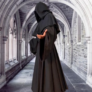 May include: A person wearing a long, black hooded robe stands in a stone hallway with arched ceilings. The robe has a wide, flowing skirt and long sleeves. The person's face is hidden by the hood.