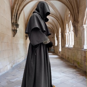 May include: A dark grey hooded robe featuring a layered shoulder cape, wide sleeves, and a floor-length skirt. The garment is constructed with a heavy fabric, providing a structured silhouette that covers the body from the head down to the feet.