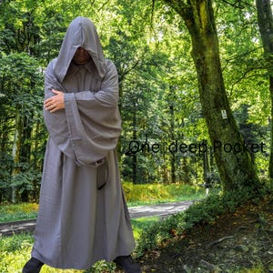 May include: A floor-length, solid grey hooded robe with long, wide sleeves and a deep pocket. The garment features a large, pointed hood that covers the head and drapes over the shoulders, with the text One deep Pocket visible on the right side.
