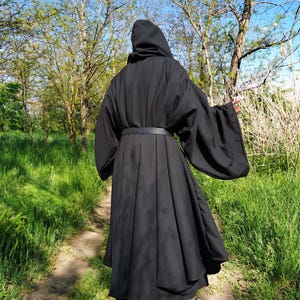 May include: A floor-length black hooded robe featuring wide, flowing sleeves and a fitted waist cinched with a matching black belt. The garment is constructed with a full, pleated skirt that drapes downwards, creating a voluminous silhouette.