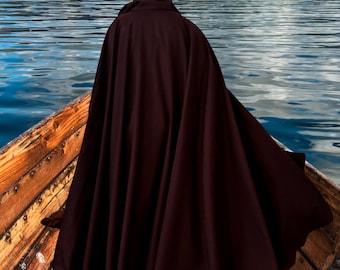 May include: A dark burgundy hooded cloak, draped over a person in a wooden boat on a lake. The cloak's fabric flows, creating a dramatic silhouette against the water and distant shoreline. The hood is up, obscuring the person's face.