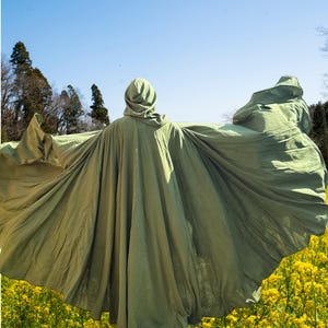 May include: A flowing, olive green hooded cloak, with the hood up, is spread wide against a backdrop of a bright blue sky and a field of yellow flowers. The cloak's fabric billows in the wind, creating a dramatic effect.
