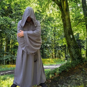 May include: A floor-length, solid grey hooded robe with long, wide sleeves and a deep pocket. The garment features a structured hood that covers the head and drapes over the shoulders, with the text One deep Pocket visible on the right side.
