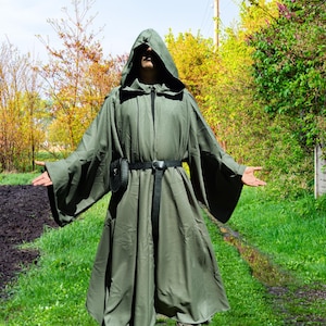 May include: A hooded olive green cloak with wide sleeves, a black belt, and a small black pouch. The cloak is worn outdoors with arms outstretched, set against a backdrop of green grass and trees.