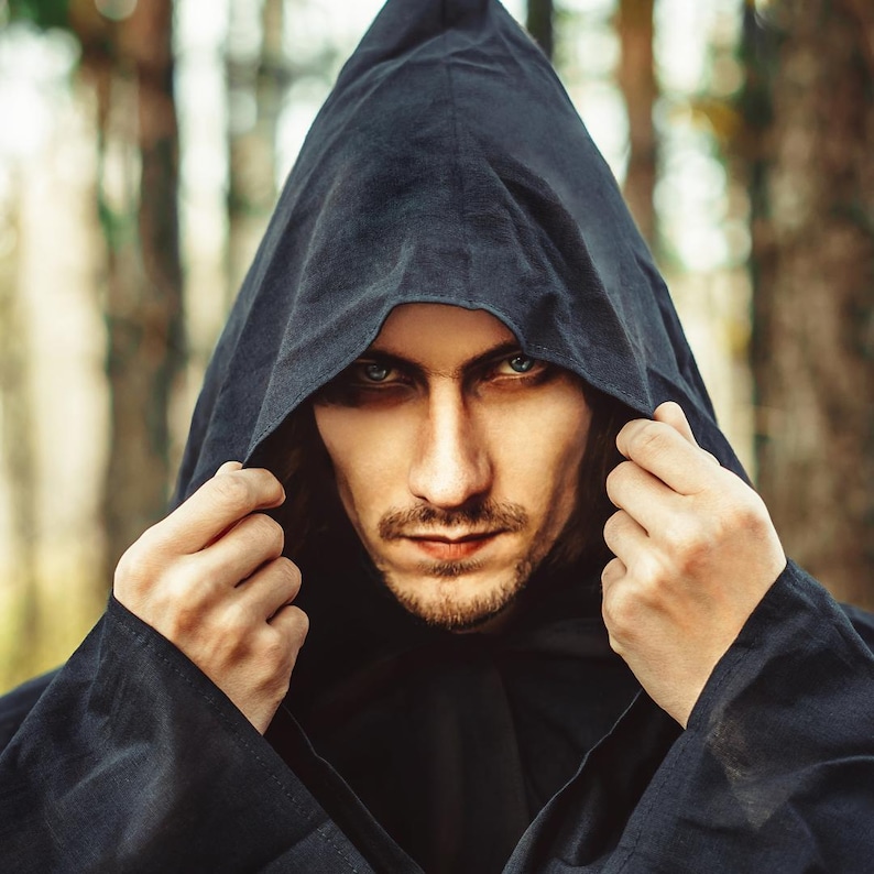 May include: A dark charcoal gray hooded cloak made of a textured fabric. The hood is pulled up over the head, framing the face with deep shadows. The garment features long sleeves and a simple, clean design with visible stitching along the hood edge.